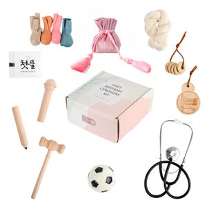 May include: A first birthday ceremony kit with a variety of items, including a wooden hammer, a stethoscope, a soccer ball, a small pouch, a wooden horse, and a card with Korean text. The kit is packaged in a pink and white box with the words "First Birthday Ceremony Kit" printed on it.
