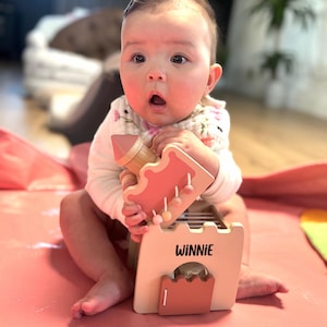 May include: A baby is holding a wooden toy castle with the name "WINNIE" printed on it. The toy is primarily peach and light brown, with a pink base. The baby is wearing a white onesie with a floral bib.