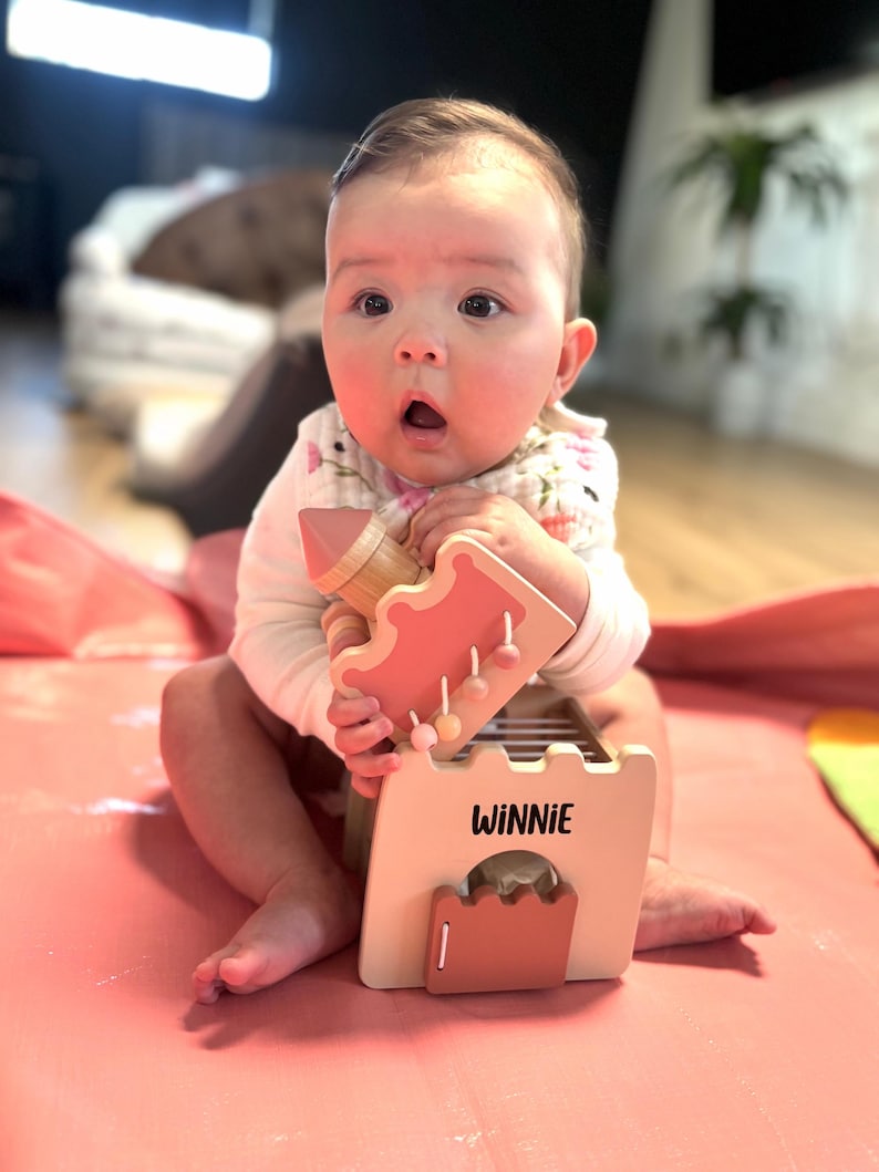 May include: A baby is holding a wooden toy castle with the name "WINNIE" printed on it. The toy is primarily peach and light brown, with a pink base. The baby is wearing a white onesie with a floral bib.