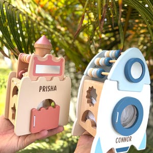 May include: Two wooden toys: a castle and a rocket. The castle is beige with pink accents and the name "PRISHA". The rocket is white and blue with the name "CONNOR". Both toys have abacus beads.