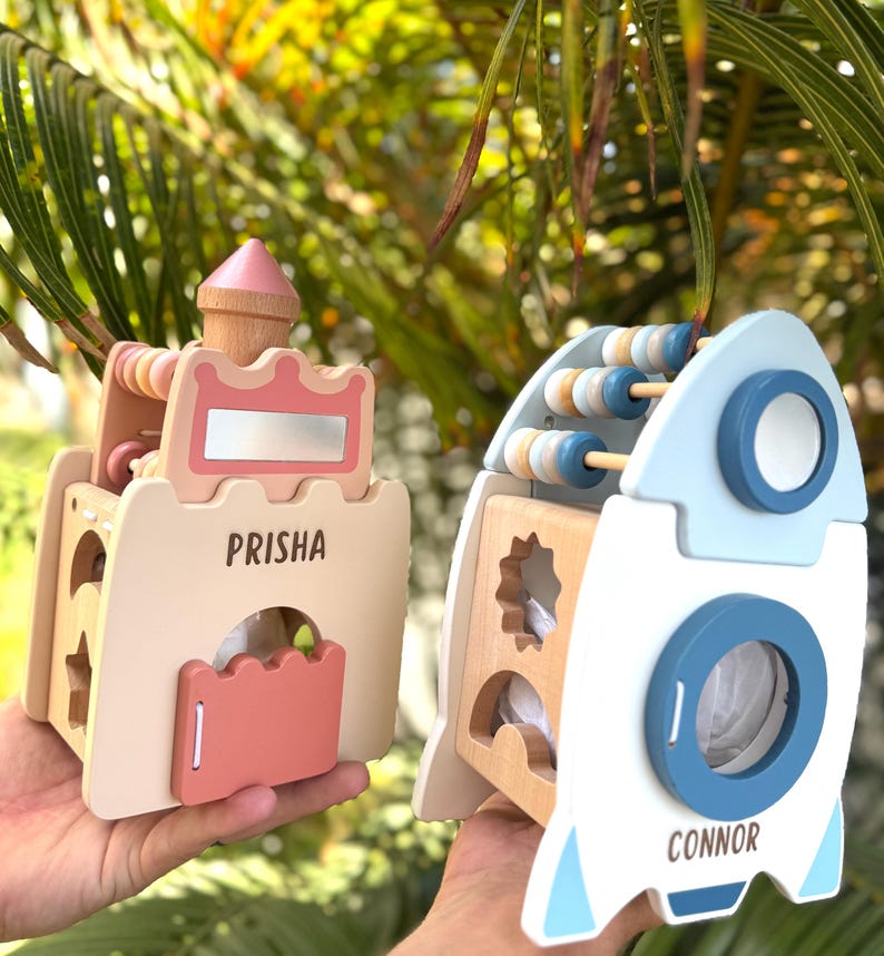 May include: Two wooden toys: a castle and a rocket. The castle is beige with pink accents and the name "PRISHA". The rocket is white and blue with the name "CONNOR". Both toys have abacus beads.