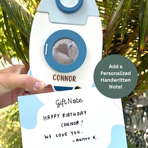May include: A white and blue rocket-shaped gift box with the name "CONNOR" on it. A handwritten gift note reads "HAPPY BIRTHDAY CONNOR! WE LOVE YOU. - Aunty K." The image also includes the text "Add a Personalized Handwritten Note!"