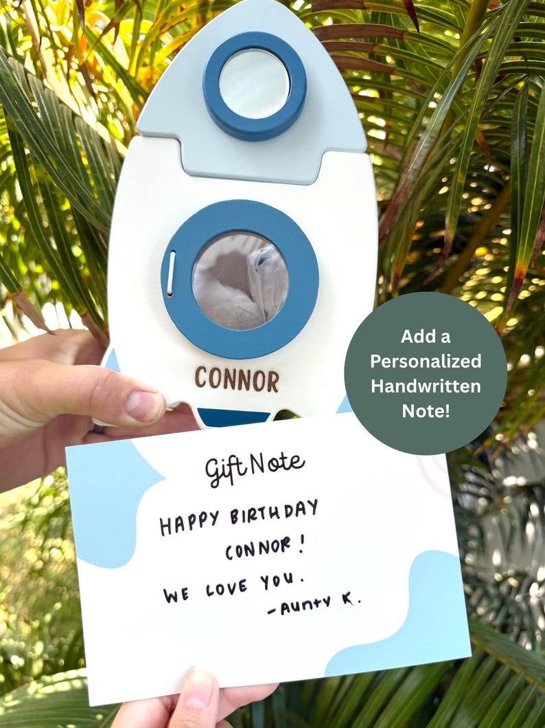 May include: A white and blue rocket-shaped gift box with the name "CONNOR" on it. A handwritten gift note reads "HAPPY BIRTHDAY CONNOR! WE LOVE YOU. - Aunty K." The image also includes the text "Add a Personalized Handwritten Note!"