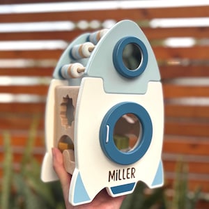 May include: A wooden rocket-shaped toy with a light blue and white color scheme. It features a bead maze, shape sorter, and a porthole with the name "MILLER" printed on the side. The toy is designed for children.