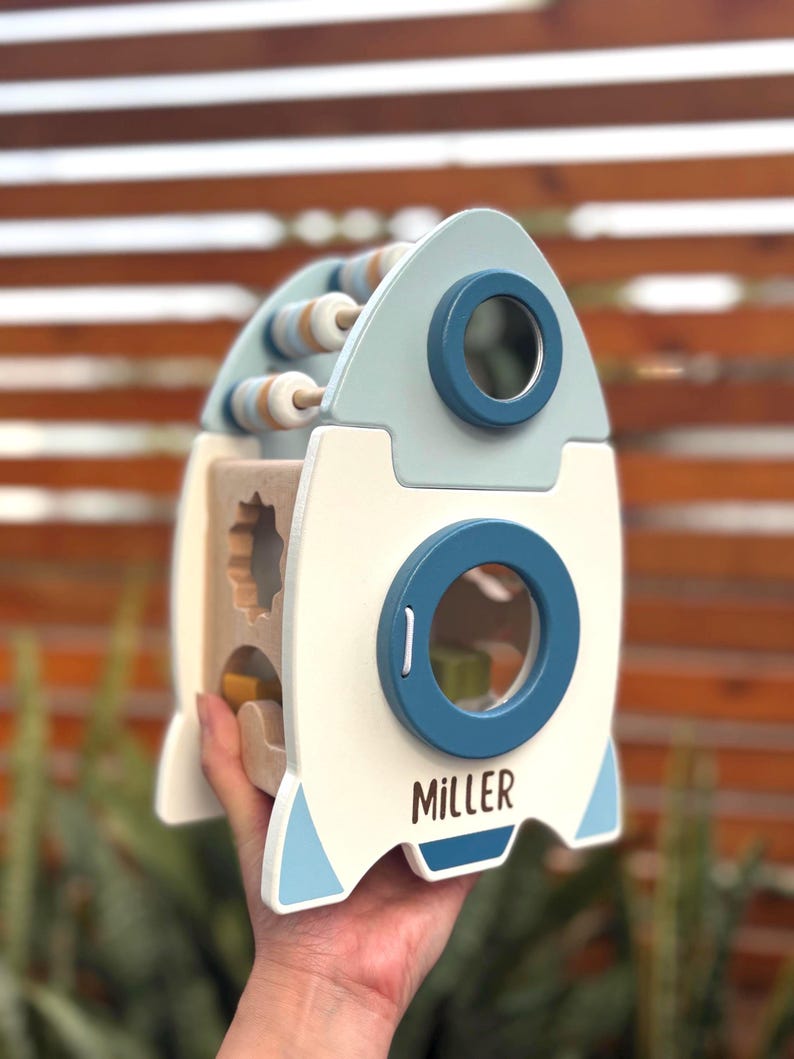 May include: A wooden rocket-shaped toy with a light blue and white color scheme. It features a bead maze, shape sorter, and a porthole with the name "MILLER" printed on the side. The toy is designed for children.
