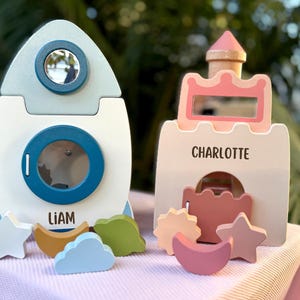 May include: Two wooden toys: a rocket ship and a castle. The rocket is white with blue accents and the name "LIAM". The castle is pink and white with the name "CHARLOTTE". Both have cut-out shapes and are on a pink surface.