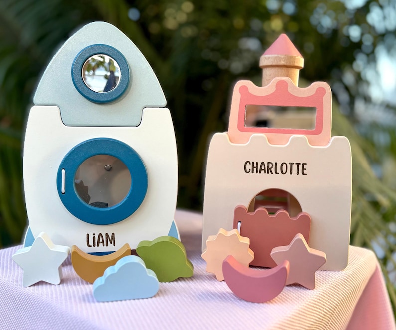 May include: Two wooden toys: a rocket ship and a castle. The rocket is white with blue accents and the name "LIAM". The castle is pink and white with the name "CHARLOTTE". Both have cut-out shapes and are on a pink surface.