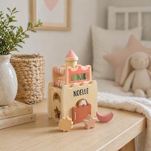 May include: Wooden toy castle with the name "NOELLE" on the front. The castle features a pink roof, a mirror, and colorful abacus beads. Includes wooden shapes: a star, a moon, and a flower. The toy is on a light wood surface.