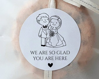 May include: Two white favor bags with a black and white illustration of a bride and groom. The bags are tied with a white ribbon and have a sticker that says "We are so glad you are here".