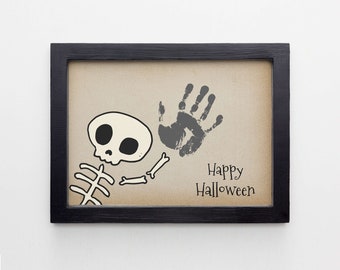 May include: A framed print with a black frame and a beige background. The print features a cartoon skeleton with a handprint in gray paint. The text "Happy Halloween" is written below the skeleton.