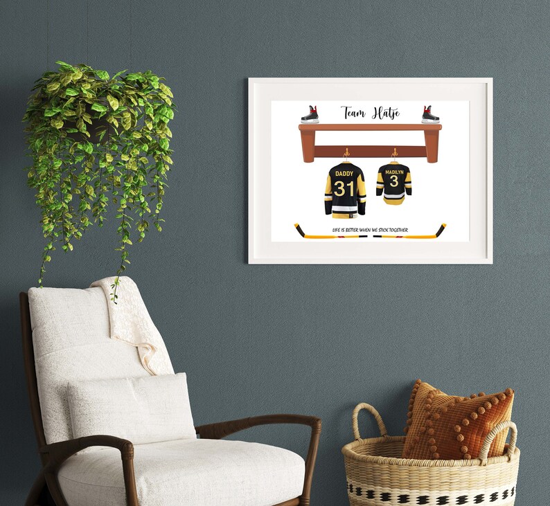 Op de afbeelding: Een wit ingelijst print met de tekst "Team Hatje" en een hockeystick met de tekst "Het leven is beter als we samen scoren". Twee hockeytruien hangen aan haken boven een houten plank met twee paar schaatsen op de plank. De truien zijn zwart met gele en witte accenten. E&eacute;n trui heeft nummer 31 en de tekst "Daddy" op de rug. De andere trui heeft nummer 3 en de tekst "Maclyn" op de rug.