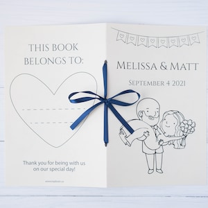 Pu&ograve; includere: Libro degli ospiti di nozze aperto con un nastro blu navy. La copertina recita "THIS BOOK BELONGS TO:" con un contorno a cuore. All'interno, presenta i nomi "MELISSA & MATT" e la data "SEPTEMBER 4 2021", insieme a un'illustrazione a cartoni animati.