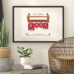Op de afbeelding: Een ingelijst print met een hockeythema. Het toont een houten plank met vier hockeytruien die eraan hangen. De truien zijn rood met witte afwerking en hebben de nummers 34, 26, 32 en 34 op de rug. De tekst "Team Richard" staat bovenaan de print. De tekst "Life is better when we stick together" staat onderaan de print.