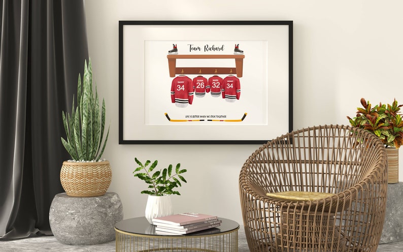 Op de afbeelding: Een ingelijst print met een hockeythema. Het toont een houten plank met vier hockeytruien die eraan hangen. De truien zijn rood met witte afwerking en hebben de nummers 34, 26, 32 en 34 op de rug. De tekst "Team Richard" staat bovenaan de print. De tekst "Life is better when we stick together" staat onderaan de print.
