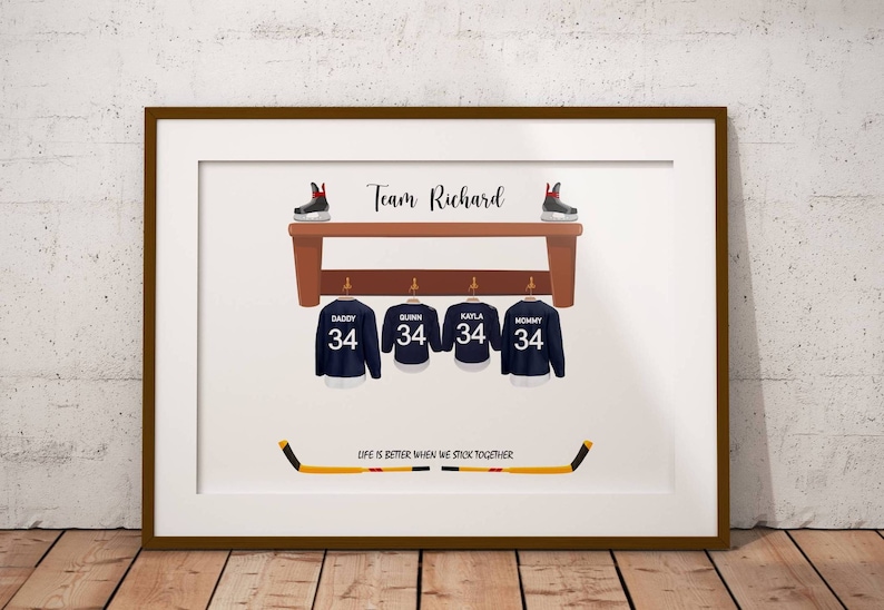 Op de afbeelding: Een witte ingelijste print met een hockeythema. De print toont vier hockeytruien die aan een houten plank hangen met de tekst "Team Richard" boven de plank. De truien hebben de namen "Daddy", "Quinn", "Kayla" en "Mommy" erop, elk met nummer 34. Onder de plank ligt een hockeystick met de tekst "Life is better when we stick together".