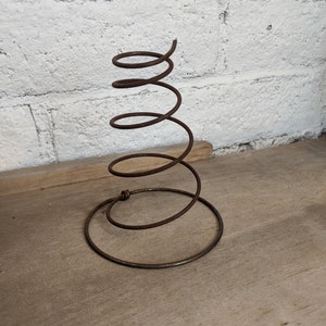 May include: A rusty metal spring, coiled and standing upright. The spring has a circular base and a spiraling upper section. It is set against a white brick wall and a wooden surface.
