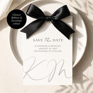 Może przedstawiać: Biała karta "Save the Date" z czarną satynową kokardą. Karta zawiera napis "SAVE the DATE", imiona Katherine & Michael, datę 10 sierpnia 2025 r. oraz "INVITATION TO FOLLOW". Inicjały "KM" znajdują się na dole. Kokarda jest czarna.