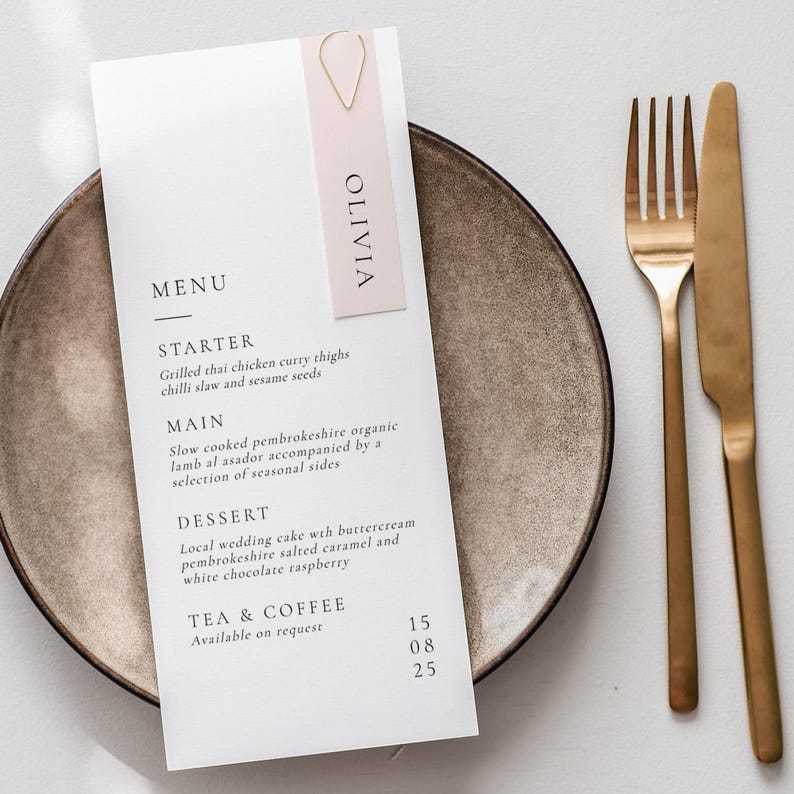 May include: A white menu card with a pink Olivia name card attached to the top. The menu lists starter, main, dessert, and tea and coffee options with prices. The menu is on a brown plate with gold cutlery.