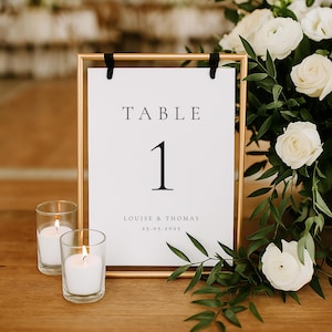 Könnte beinhalten: Ein goldfarbener Tischnummernschild mit dem Text "TABLE 1" und den Namen "LOUISE & THOMAS" sowie dem Datum "25.05.2025".
