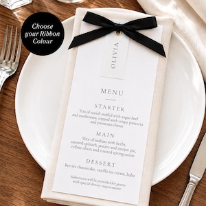 May include: A white rectangular menu card with black text listing a three-course meal, featuring a name tag labeled OLIVIA attached with a black satin ribbon bow. The menu is placed on a folded beige napkin atop a white plate with a fork and knife.