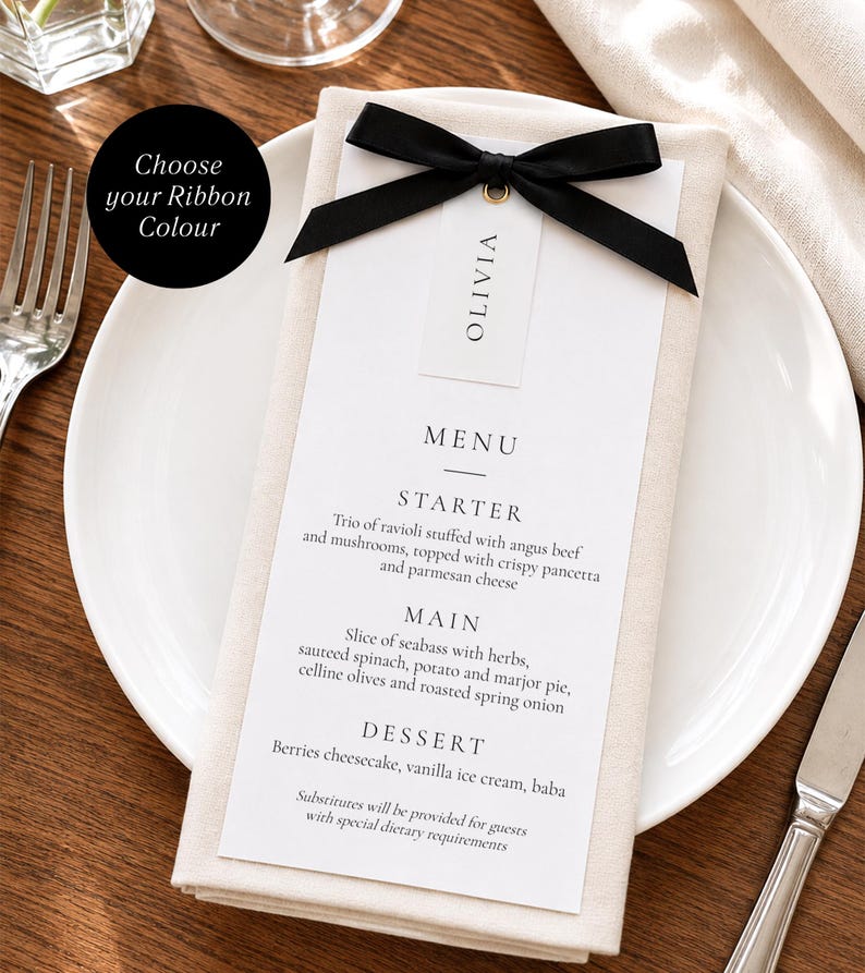 May include: A white rectangular menu card with black text listing a three-course meal, featuring a name tag labeled OLIVIA attached with a black satin ribbon bow. The menu is placed on a folded beige napkin atop a white plate with a fork and knife.