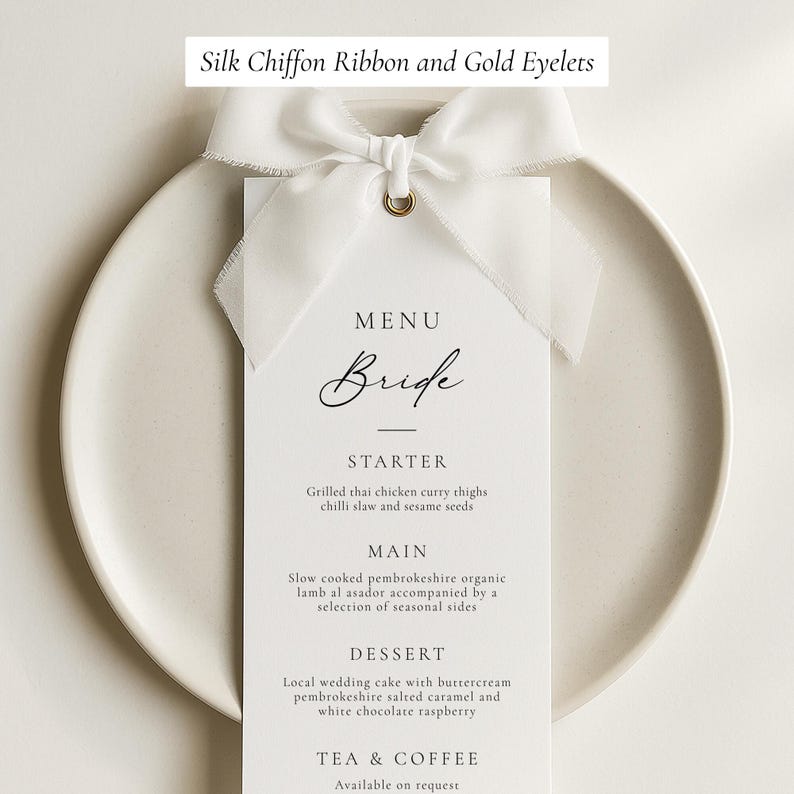 May include: A white menu card with the word "Bride" in elegant script, tied with a silk chiffon ribbon and gold eyelet. The menu lists starter, main, and dessert options, including grilled Thai chicken and wedding cake.