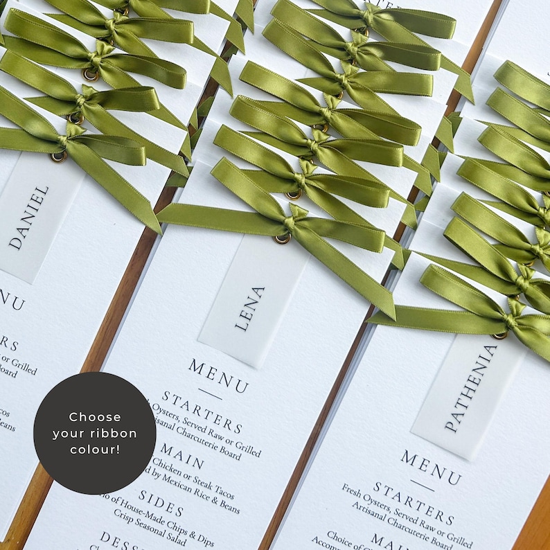 May include: White place cards with olive green satin ribbon bows. Each card has a name printed on it. The cards are part of a menu with the text "MENU", "STARTERS", "MAIN", "SIDES", and "DESSERTS". A circular graphic says "Choose your ribbon colour!"