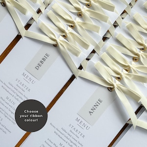 May include: Elegant wedding menu cards with ivory ribbon bows. Each card features a guest's name and a detailed menu, including starters and main courses. The cards are arranged on a wooden surface, creating a sophisticated presentation.