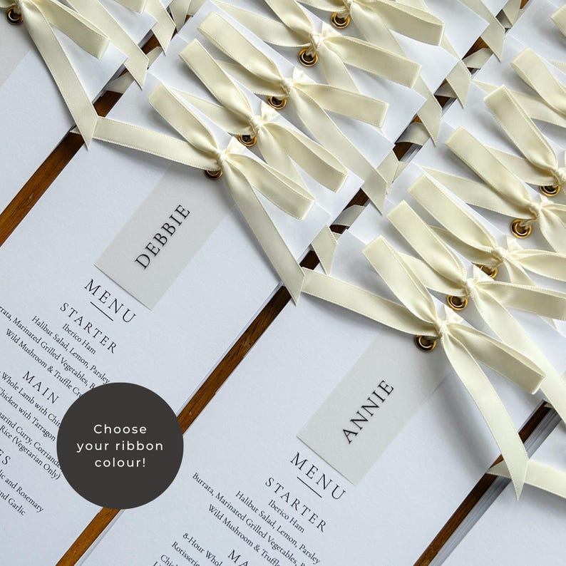 May include: Elegant wedding menu cards with ivory ribbon bows. Each card features a guest's name and a detailed menu, including starters and main courses. The cards are arranged on a wooden surface, creating a sophisticated presentation.