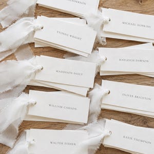May include: A collection of rectangular white paper place cards, each featuring a name printed in black serif font. Every card is attached to a frayed white silk ribbon through a small hole, displaying names such as Melissa Henderson, Michael Hopkins, Yvonne Wright, Hayleigh Dawson, Maddison Daily, Oliver Brighton, William Carson, and Katie Thompson.