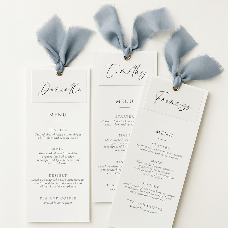 Op de afbeelding: Drie verticale menukaarten van wit papier met zwarte tekst, elk voorzien van een naamkaartje bovenaan met de namen Danielle, Timothy of Franciss. Elk menu is vastgebonden met een stoffig blauw gerafeld zijden lint door een gouden metalen oogje.