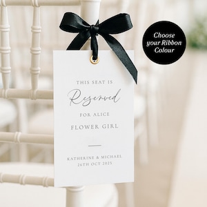 Op de afbeelding: Een witte, rechthoekige trouwstoelreserveringstag met een gouden oogje, zwart lint en tekst die luidt: "THIS SEAT IS RESERVED FOR ALICE FLOWER GIRL". De tag is vastgemaakt aan een witte stoel. Een zwarte cirkel met de tekst "Choose your Ribbon Colour" staat rechts.