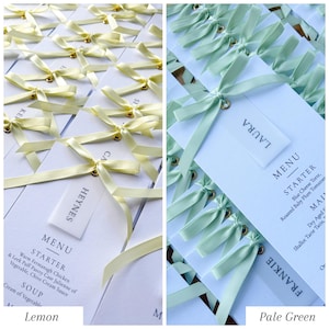 May include: Arrangement of wedding menu cards with pale yellow and pale green ribbons. Each card has a small tag with a name and a menu with starter and soup options. The cards are tied with a ribbon and a gold eyelet.
