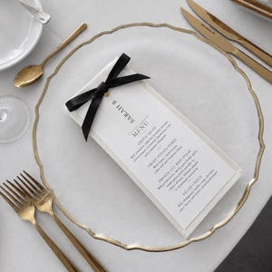 May include: A white rectangular menu card titled SARAH B MENU, featuring a black satin ribbon bow at the top, placed on a clear glass charger plate with a gold rim. The menu lists Exotic Salad, Sweet Potato Puree, French Cut Beef Steak, and Filled Choux.