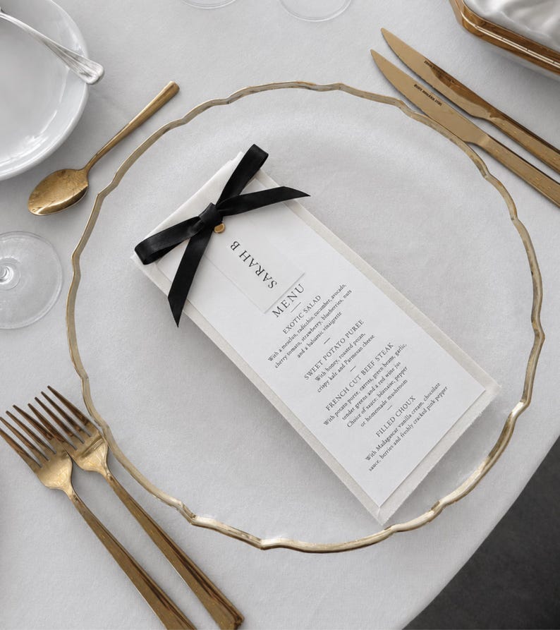 May include: A white rectangular menu card titled SARAH B MENU, featuring a black satin ribbon bow at the top, placed on a clear glass charger plate with a gold rim. The menu lists Exotic Salad, Sweet Potato Puree, French Cut Beef Steak, and Filled Choux.
