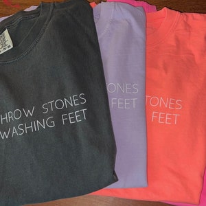 May include: Five t-shirts with different colors and the same text printed on them. The text reads "Can't throw stones while washing feet" on a dark gray shirt, "Stones feet" on a light purple shirt, "Stones feet" on a coral shirt, and "Stones feet" on a pink shirt.