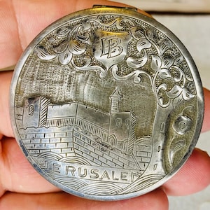 May include: Silver-toned compact with an engraved design of a building with a tree and the word "JERUSALEM". The compact has a monogram "IB" on the top.