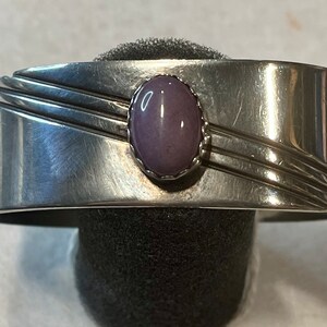 May include: A silver cuff bracelet with a purple oval gemstone set in the center. The bracelet has a simple design with three lines running across the top.
