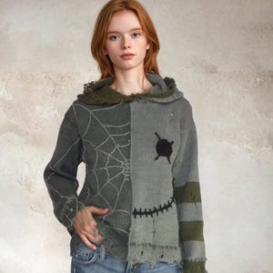 May include: A gray and green sweater with a unique design. One side features a spiderweb pattern, while the other has a stitched-together look with a black patch and a stitched smile. The sweater has a frayed hem and cuffs, and a green collar.