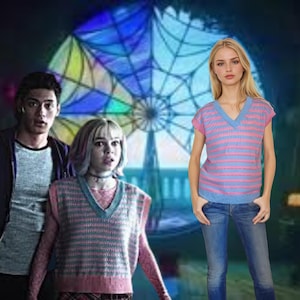 May include: A woman in a pink and blue striped V-neck sweater and blue jeans. Two other people are in the background. The sweater has a light blue V-neck and pink and blue horizontal stripes.