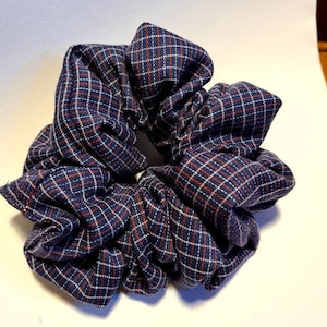 May include: A navy blue scrunchie with a red and white plaid design. The fabric is gathered and knotted, forming a voluminous hair accessory. This scrunchie is a fashionable and functional hair accessory.