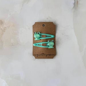 May include: Two light green hair clips with frog designs are displayed on a brown tag. The clips are made of metal and plastic. The tag has the words "caquette passion" written on it.
