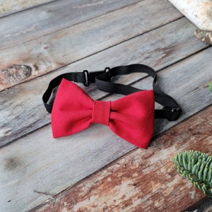 May include: A red bow tie with an adjustable black elastic strap. The bow tie is made of a soft, textured fabric.