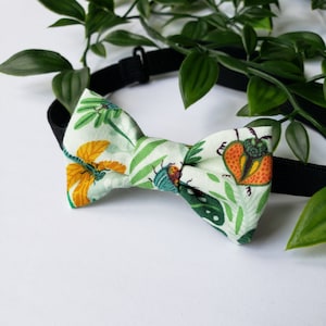 May include: A green, yellow and orange bow tie with a bug and flower pattern. The bow tie is made of fabric and has an adjustable black strap.