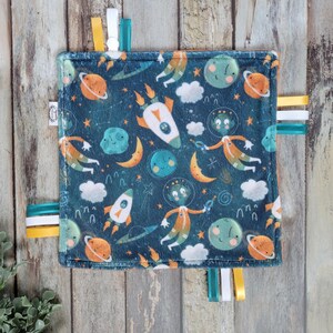 May include: A square, dark teal baby comfort blanket with a space-themed pattern featuring rockets, planets, aliens, and clouds. The blanket has colourful ribbons in yellow, white, and teal around the edges.