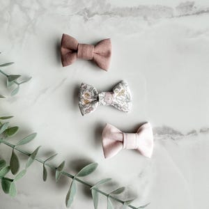 May include: Three fabric bow ties in shades of pink and floral patterns are arranged on a marble surface. The top and bottom bows are solid pink, while the centre bow features a delicate floral print. A sprig of green leaves is in the lower left corner.