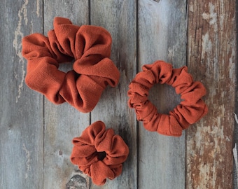 May include: Three rust-coloured fabric hair scrunchies of varying sizes. Each scrunchie features a gathered, pleated design made from a solid-coloured textile material, arranged in a cluster to display the different diameters of the hair accessories.
