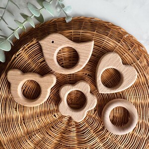 May include: Four wooden teethers in the shapes of a bird, a flower, a heart, and a bear. The teethers are on a woven wicker surface.