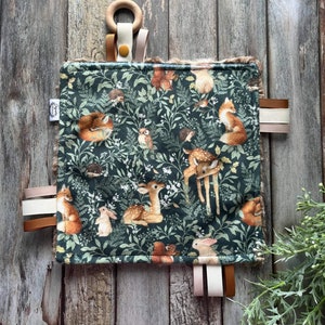 Peut inclure: Une couverture pour bébé de couleur verte avec un imprimé d'animaux de la forêt. La couverture est dotée d'un anneau en bois et de rubans de satin attachés aux coins.