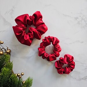 May include: Three red satin scrunchies of different sizes. The largest scrunchie is in the top left corner, the medium scrunchie is in the centre, and the smallest scrunchie is in the bottom right corner.